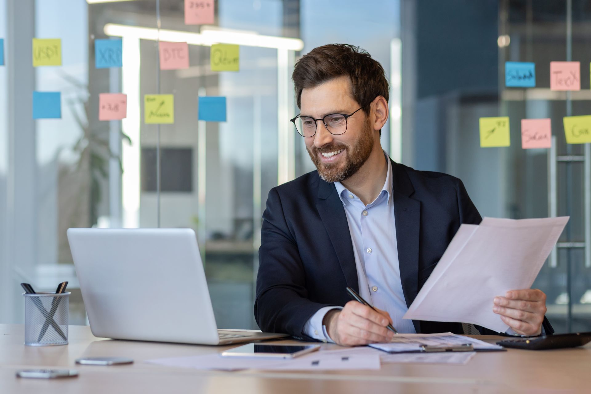 Man reviewing documents at a desk with a laptop in a bright office, surrounded by sticky notes on a glass wall