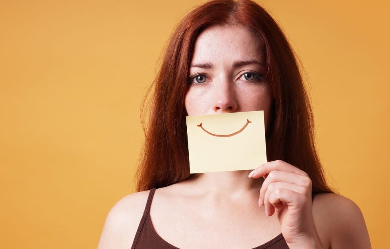 Person holding a smiling sticky note over their mouth against a yellow background