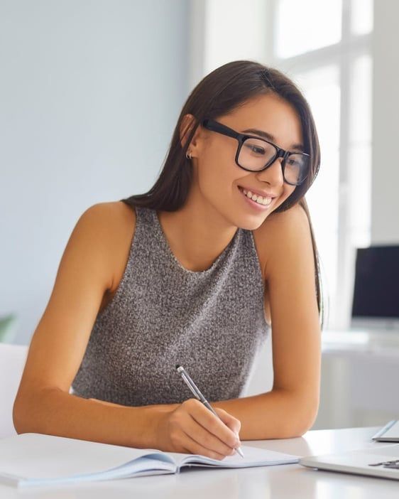 Woman in glasses writing in a notebook at a desk, smiling in a bright office.