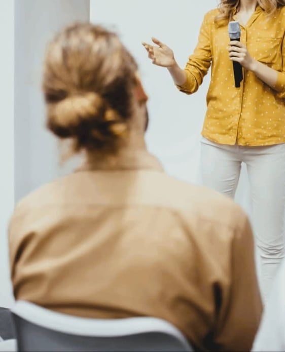 Person speaking to seated audience member in a room, holding a microphone and gesturing during a presentation