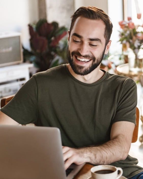 Smiling man using a laptop at a table with a cup of coffee in a bright home office