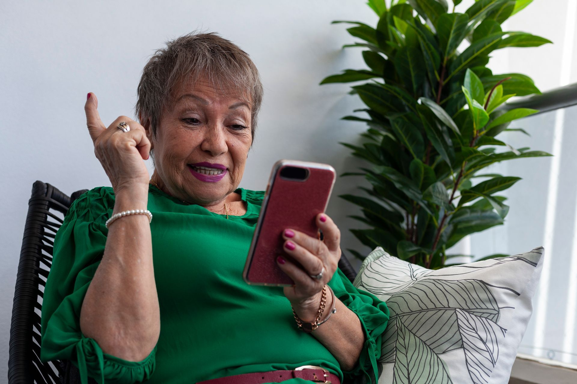 Person in a green top smiling at a smartphone beside a leafy plant on a couch
