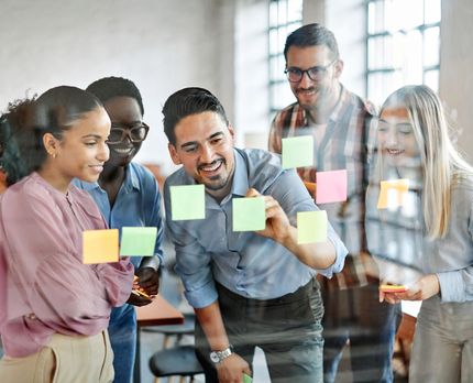 Presenter in a meeting room explains a whiteboard plan to seated colleagues.