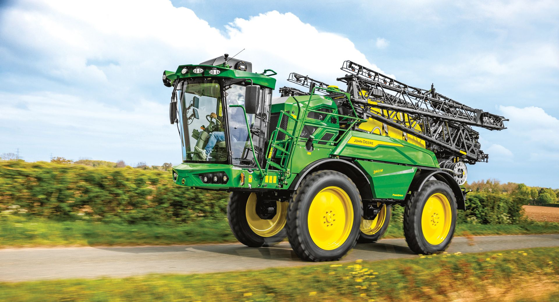 Green John Deere self-propelled sprayer on a road, spraying farm fields, yellow wheels.