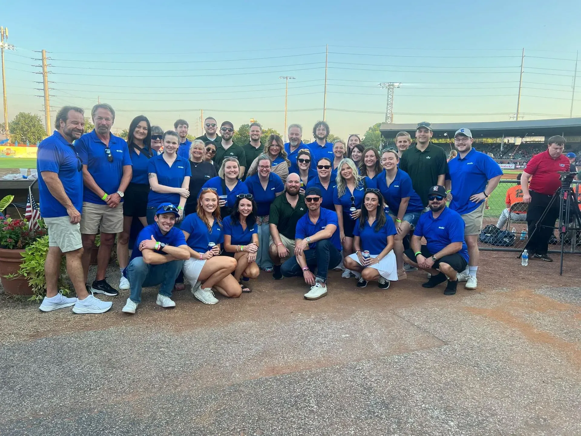Group of people in blue shirts posing outdoors on gravel at dusk, with a track field and lights behind them