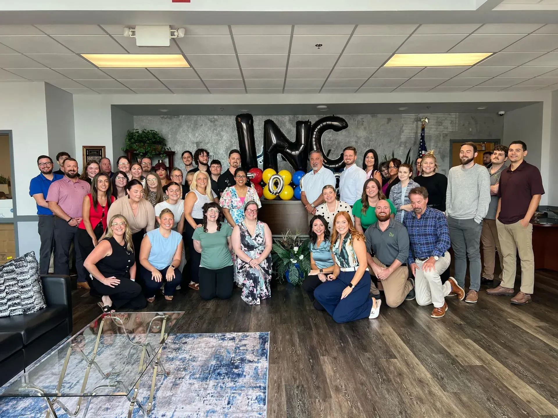 Large group posing indoors in a lobby, with balloons and a “NC” sign in the background.