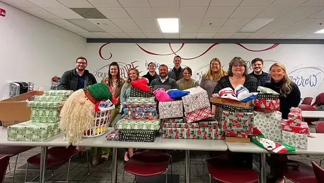 Group of people standing behind a table filled with wrapped gifts in a decorated room