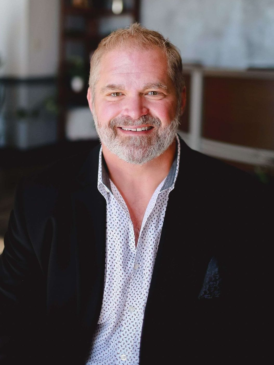 Smiling man in a dark blazer and patterned shirt, seated indoors against a softly blurred background