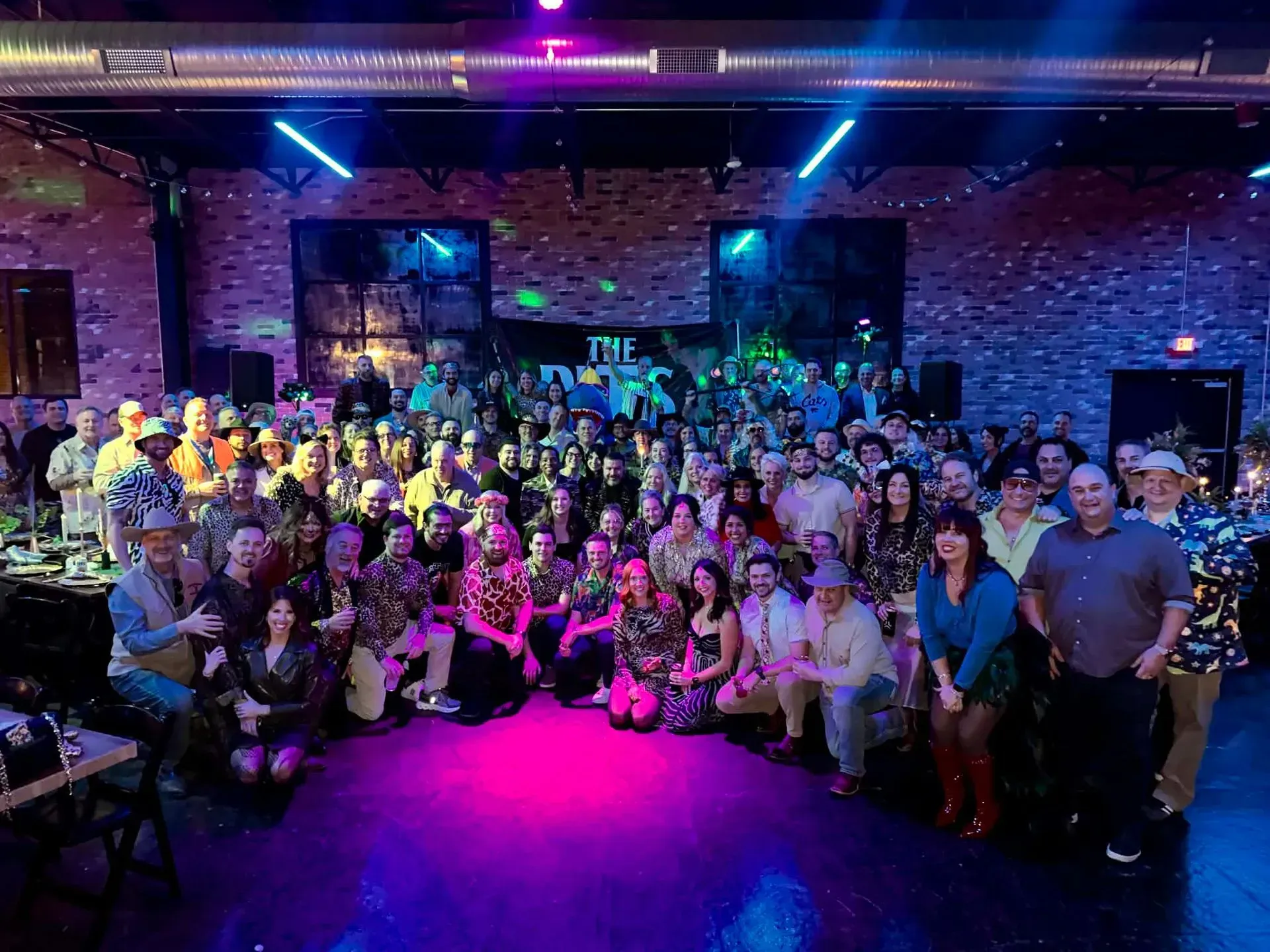 Large group posing together at a nightclub with purple and blue lights and a brick wall backdrop