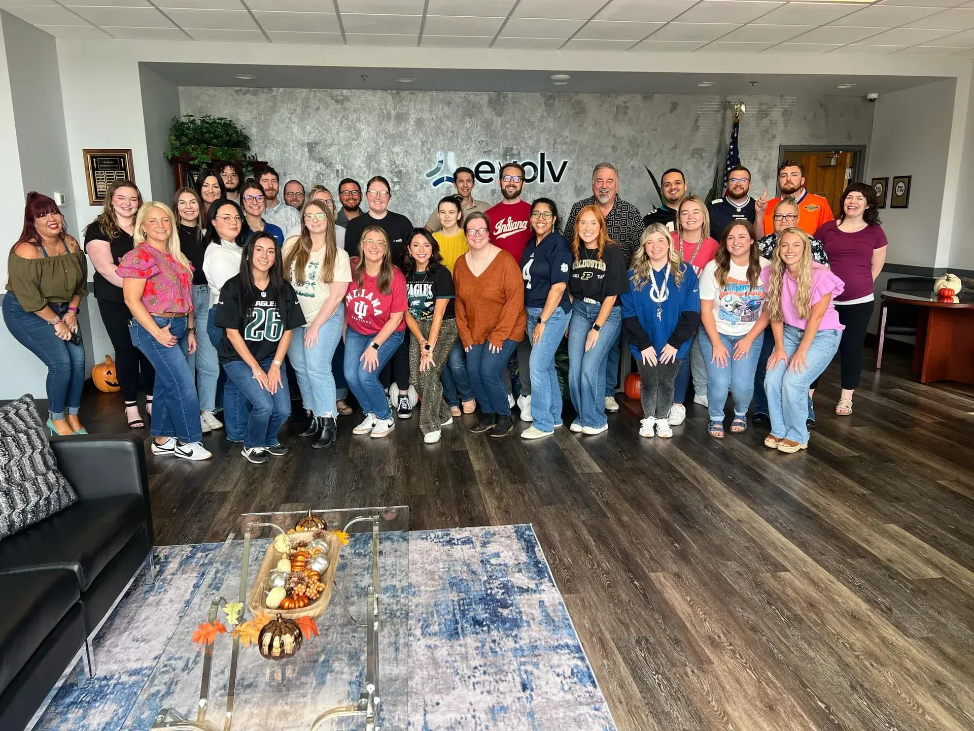 Group of people posing in a lobby, seated and standing in colorful shirts behind a patterned rug and coffee table.