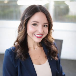 Smiling woman in a navy blazer standing indoors against a bright blurred background