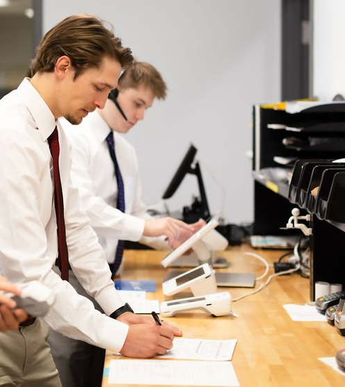 Two workers in white shirts and ties reviewing documents at a desk with computer monitors and equipment