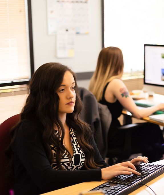 Woman typing at a desktop computer in an office, with another person working in the background.