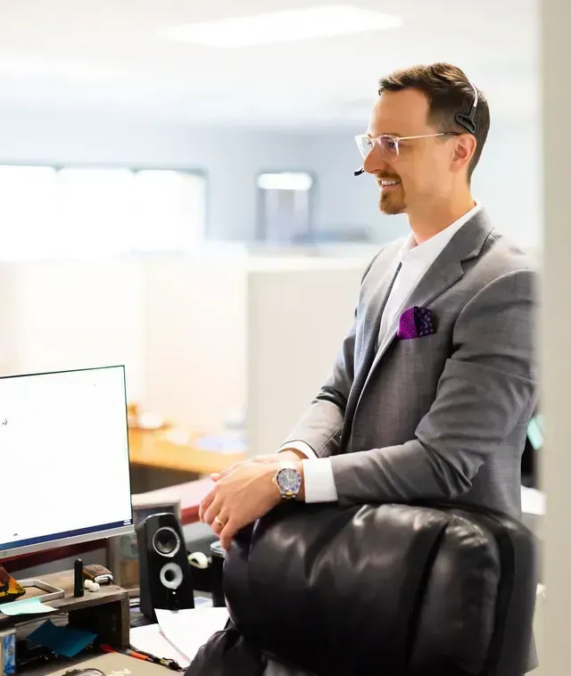 Man in a gray suit with purple pocket square stands by a desk in a bright office, smiling at his computer