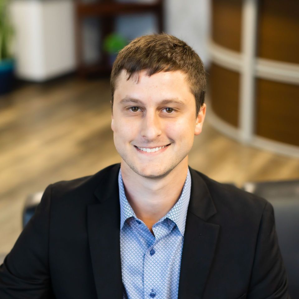 Smiling person in a dark blazer and blue shirt seated indoors with a blurred office-style background