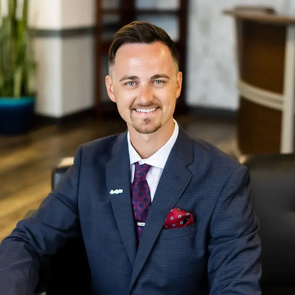 Smiling man in navy suit with purple tie and red pocket square, seated indoors.