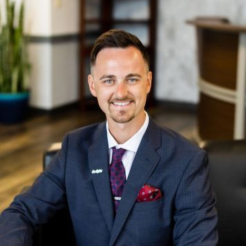 Smiling man in a navy suit with a purple tie and red pocket square, seated indoors