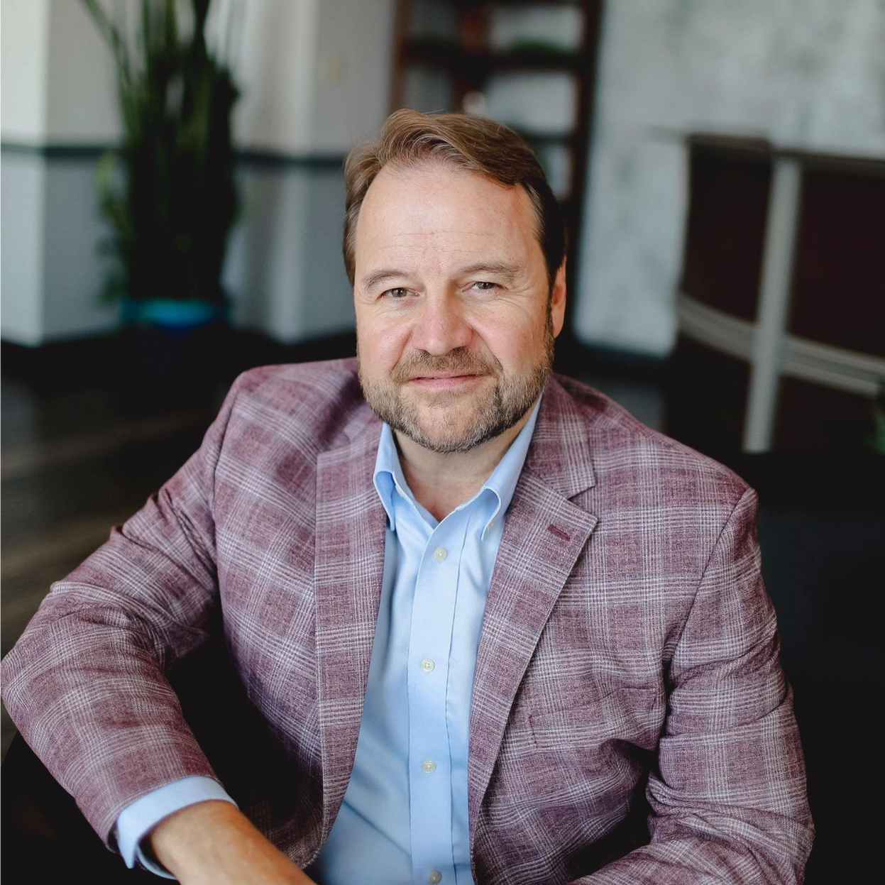 Man seated indoors in a pink plaid blazer, looking at the camera with a slight smile.