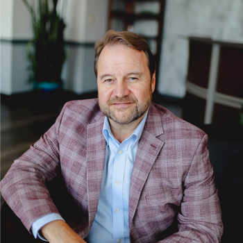 Man in a pink plaid blazer and blue shirt sitting indoors, looking at the camera