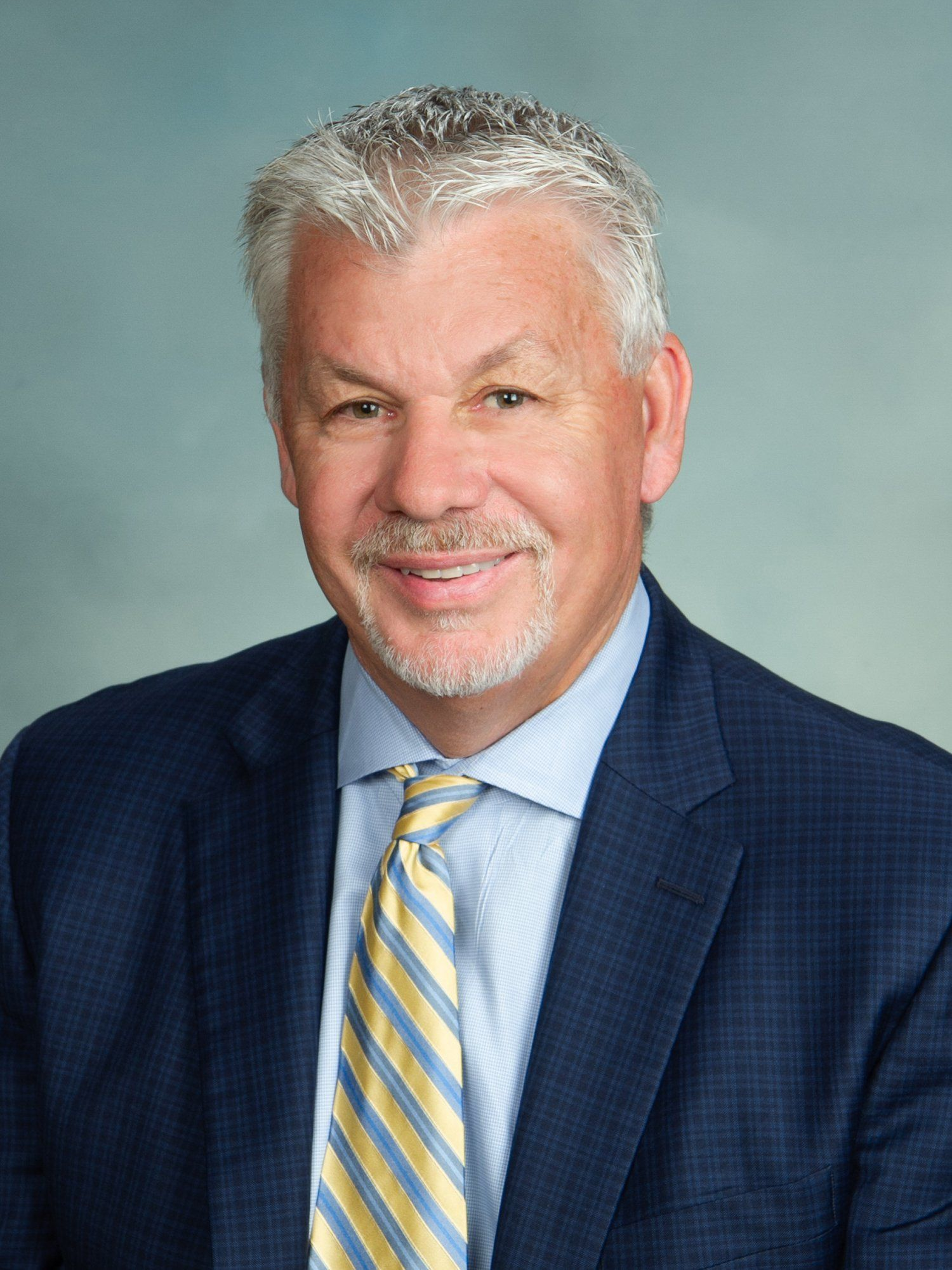 Studio headshot of a smiling man in a navy suit, striped tie, on a light blue background