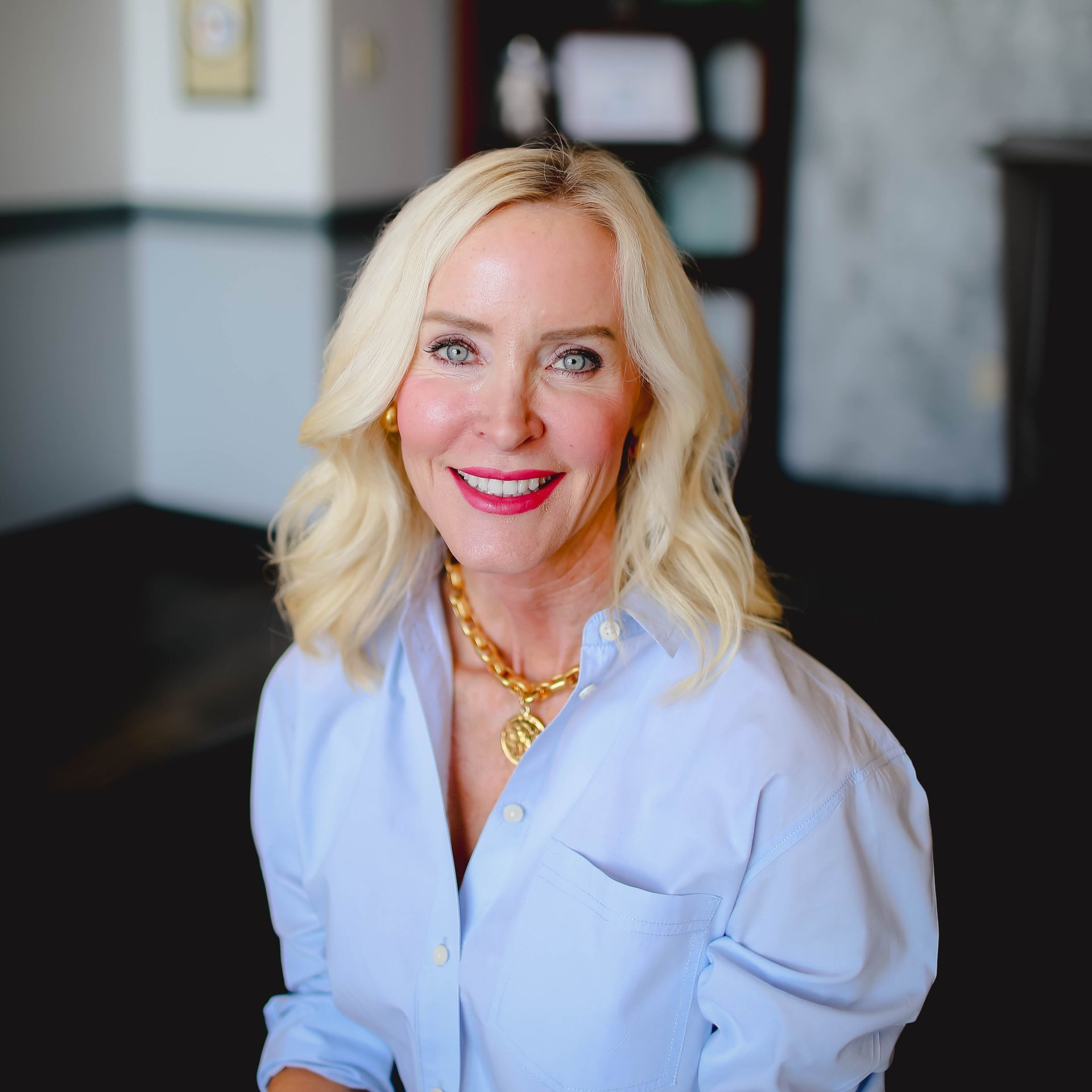 Smiling blonde woman in a light blue blouse and gold necklace in a softly blurred indoor setting