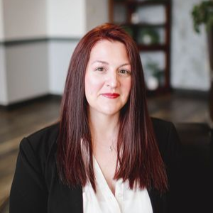 Woman with long red hair in a black blazer and white blouse, smiling in an office setting