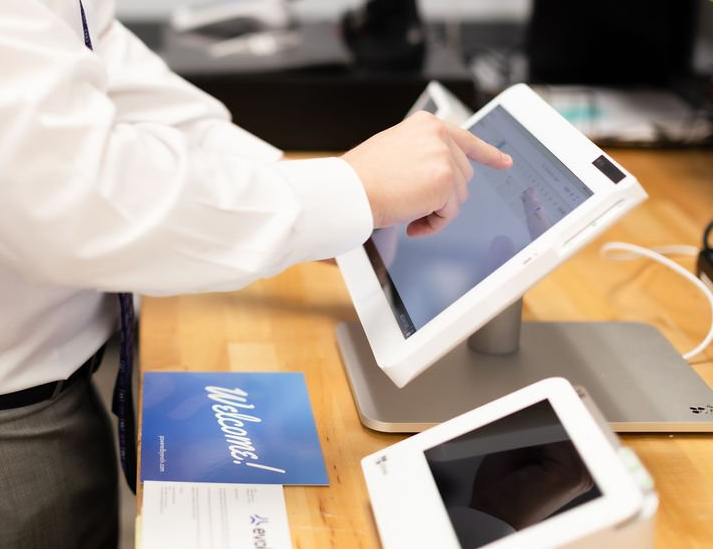 Person tapping a white tablet on a wooden table beside another tablet and a blue brochure.