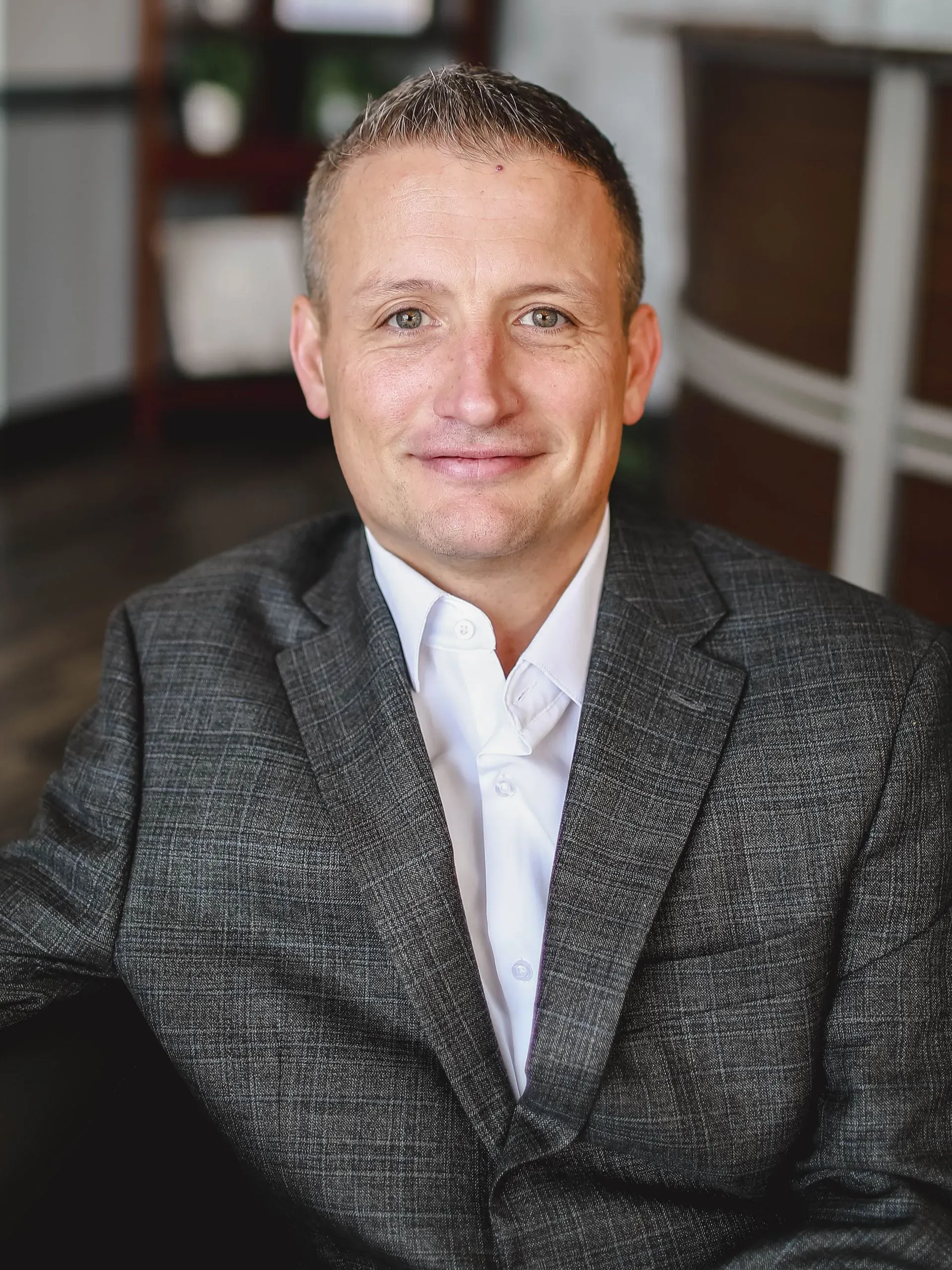Smiling man in a gray blazer and white shirt, seated indoors with a blurred background.