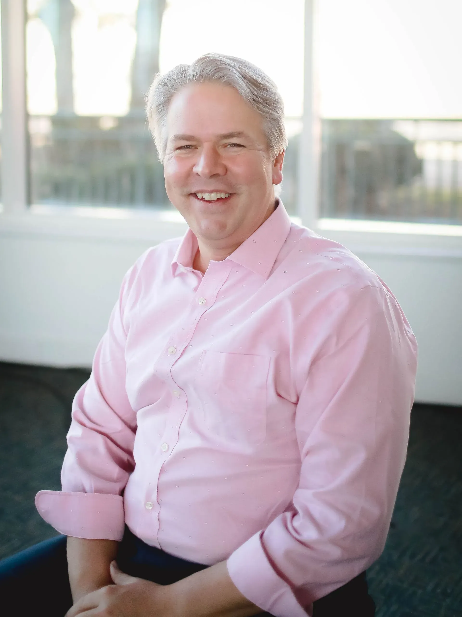 Man in a pink shirt smiling while seated indoors near a bright window