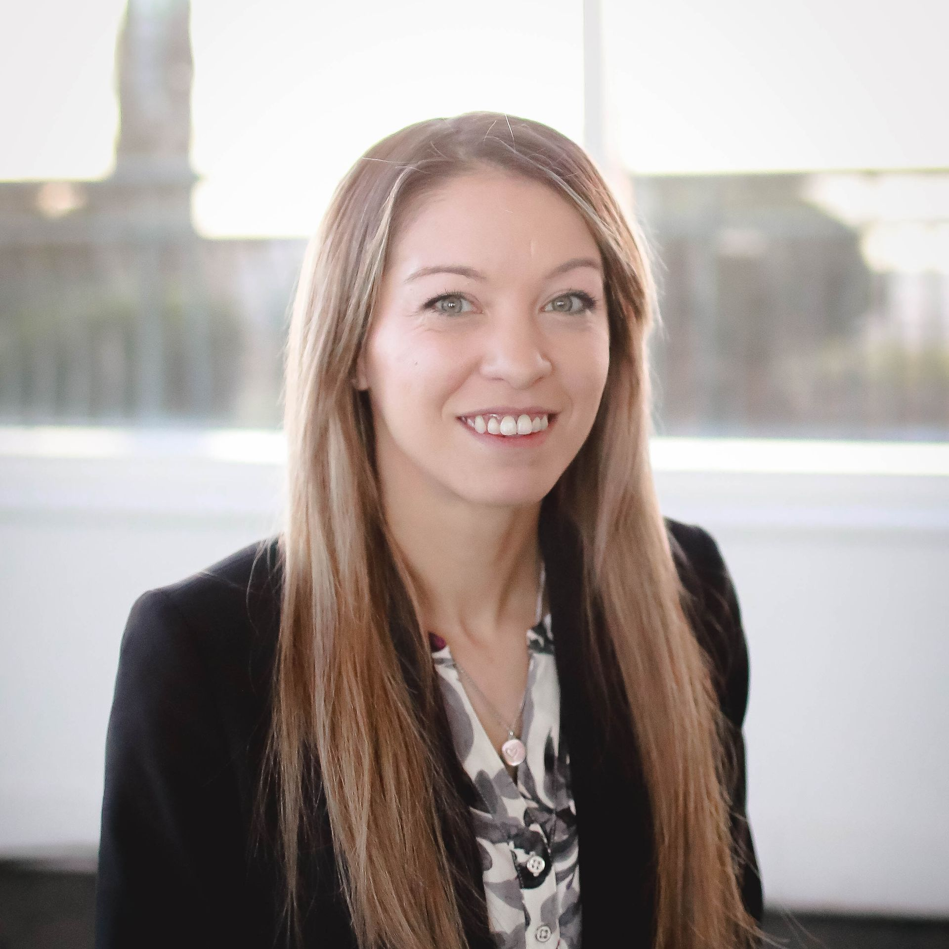 Smiling woman in a black blazer standing indoors near a bright window