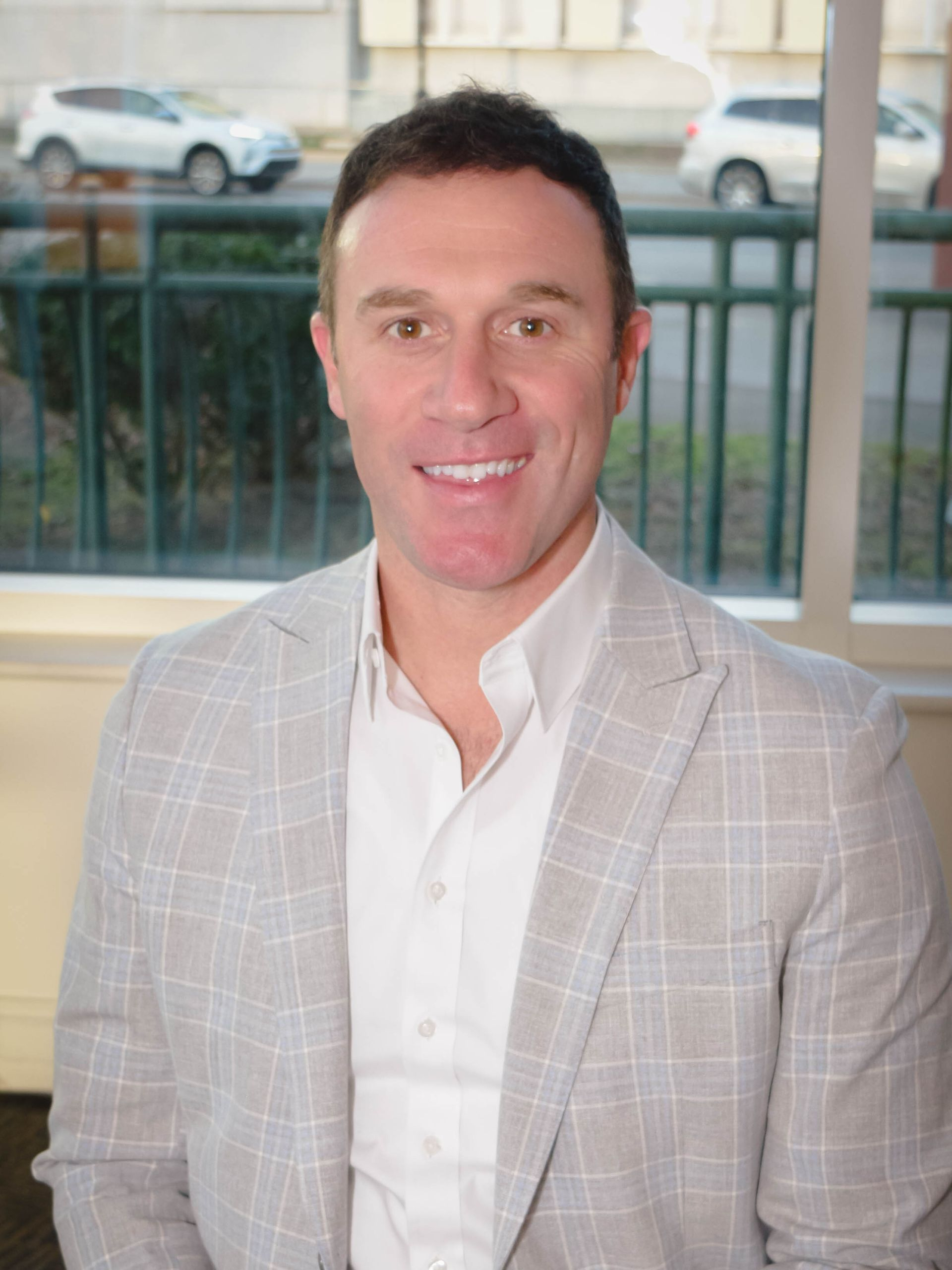 Man in a light gray plaid blazer and white shirt, smiling indoors near a window.