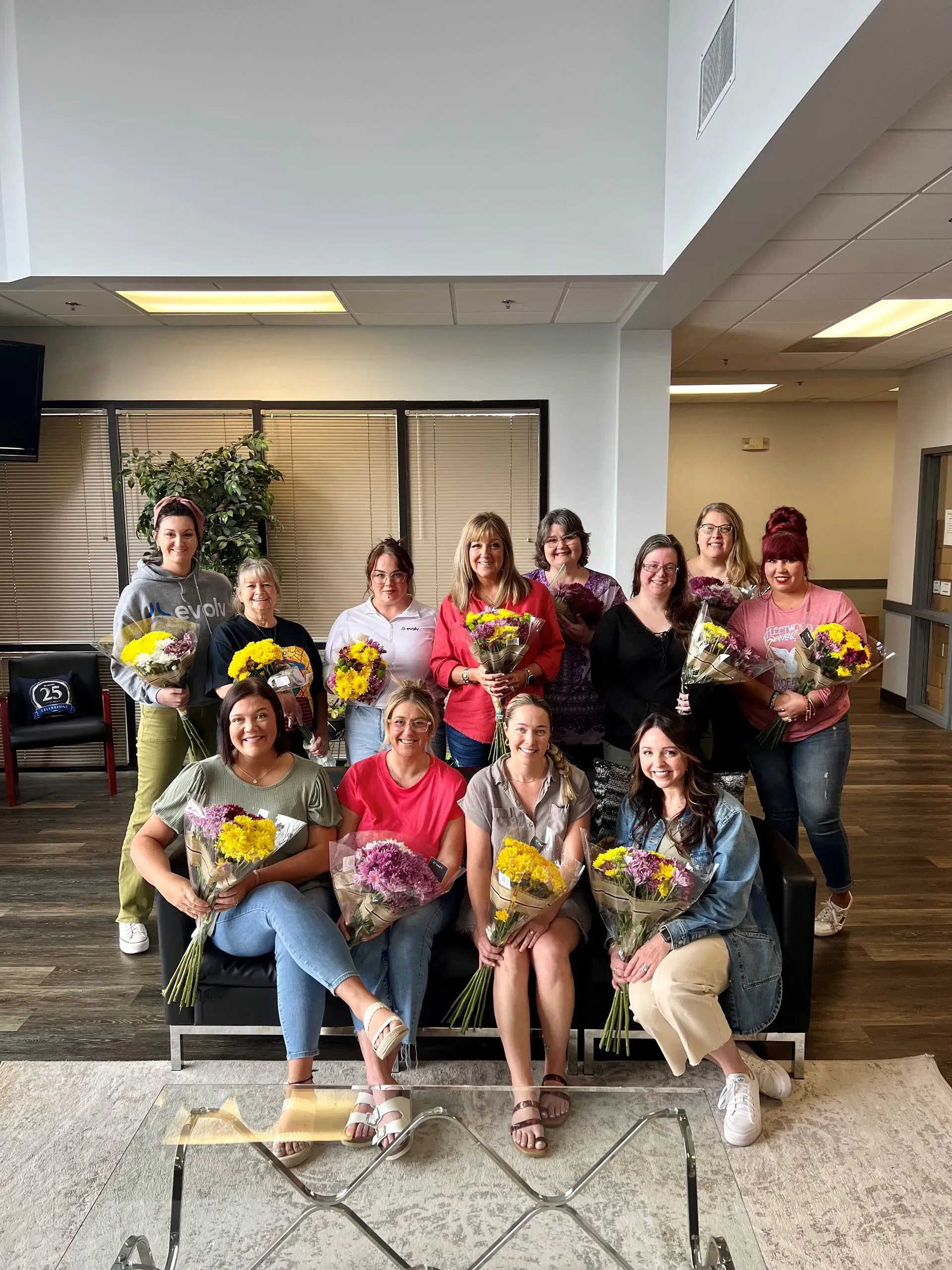 Group of people posing indoors on steps, holding colorful bouquets and smiling for a celebration photo