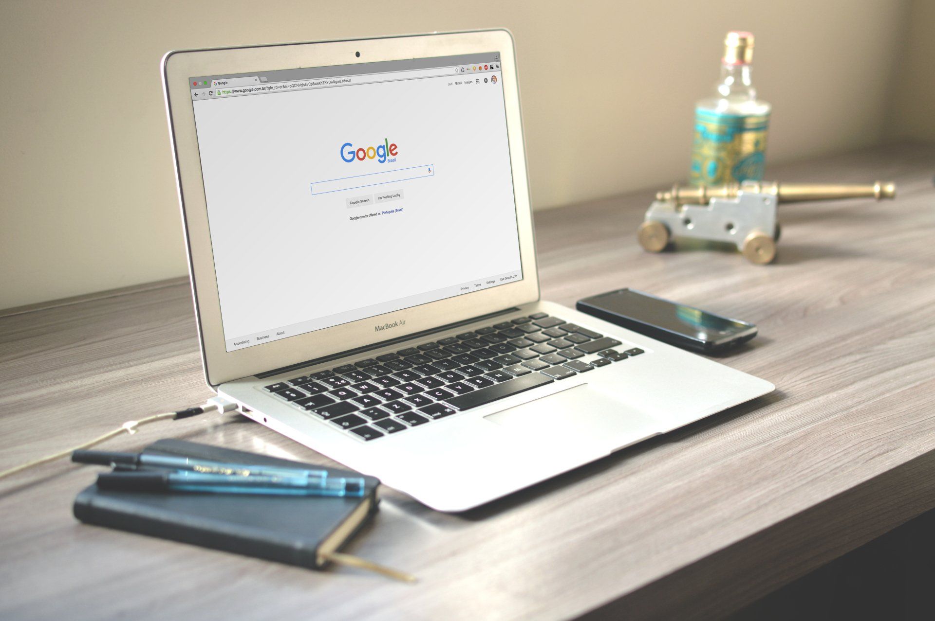 Laptop on desk displaying Google, with a smartphone and pen in the foreground and a small robot toy in back