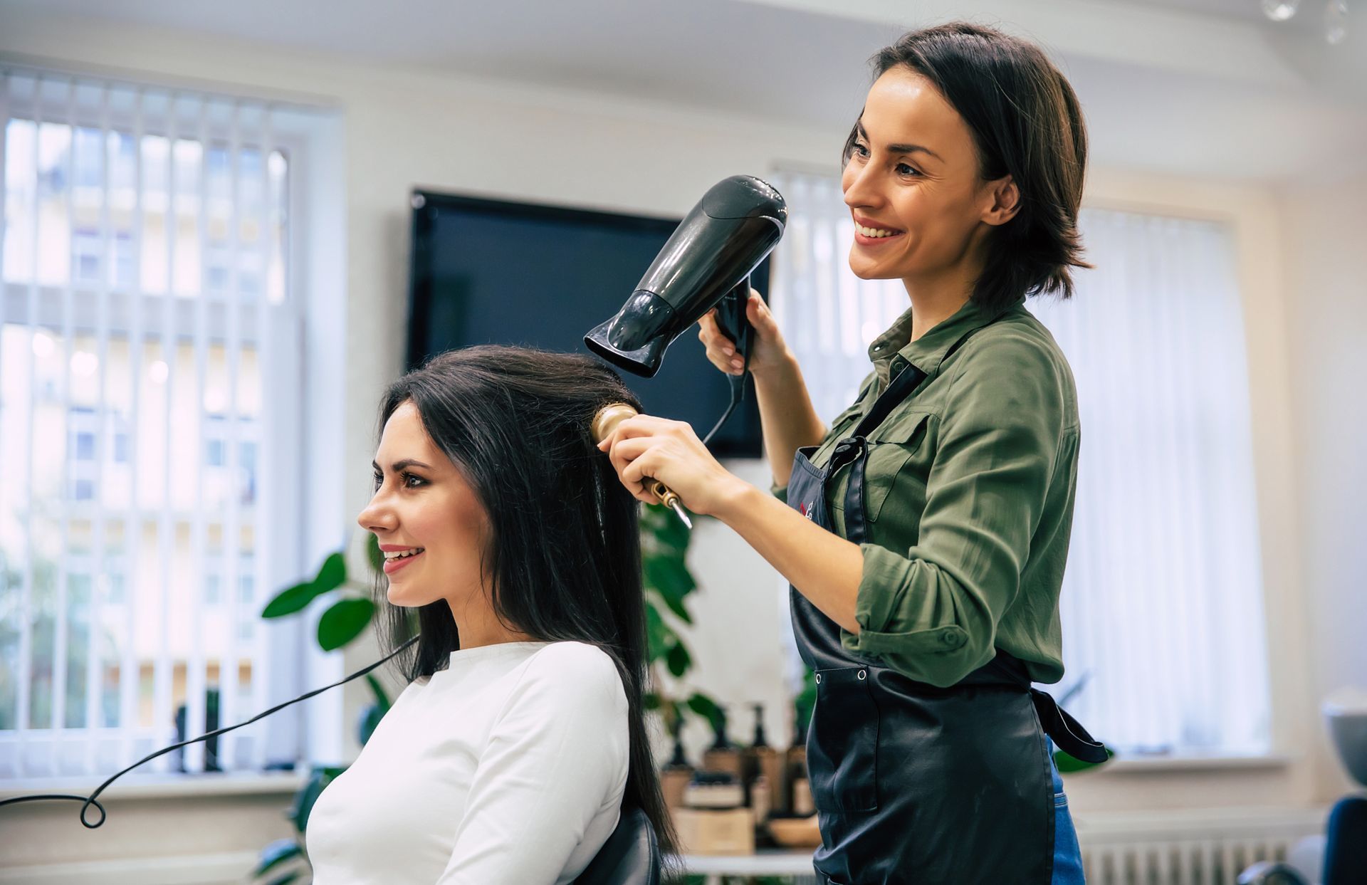 Hair stylist blow-drying a smiling client in a bright salon