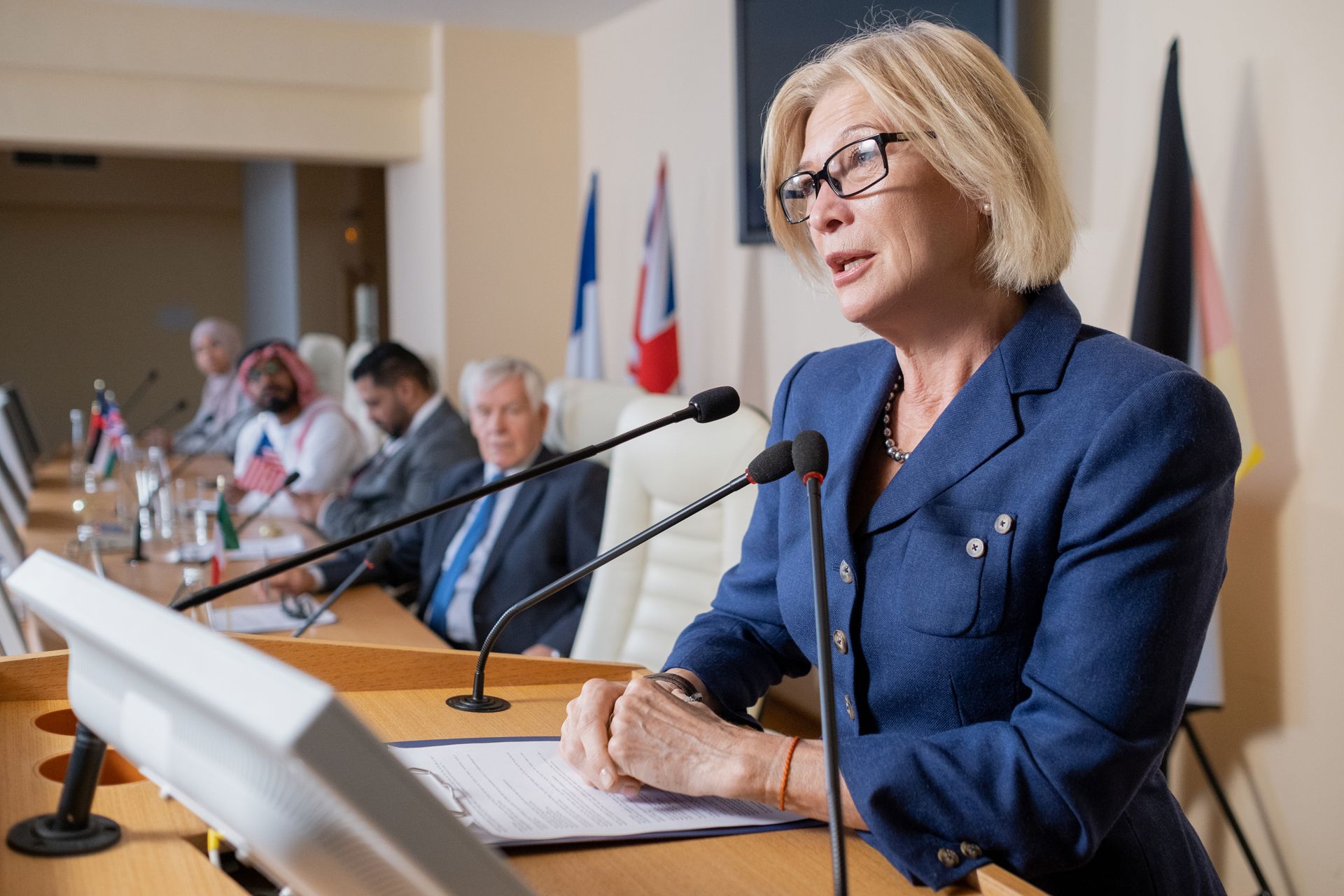 Woman speaking at a podium in a conference room, with microphones and seated attendees in the background.