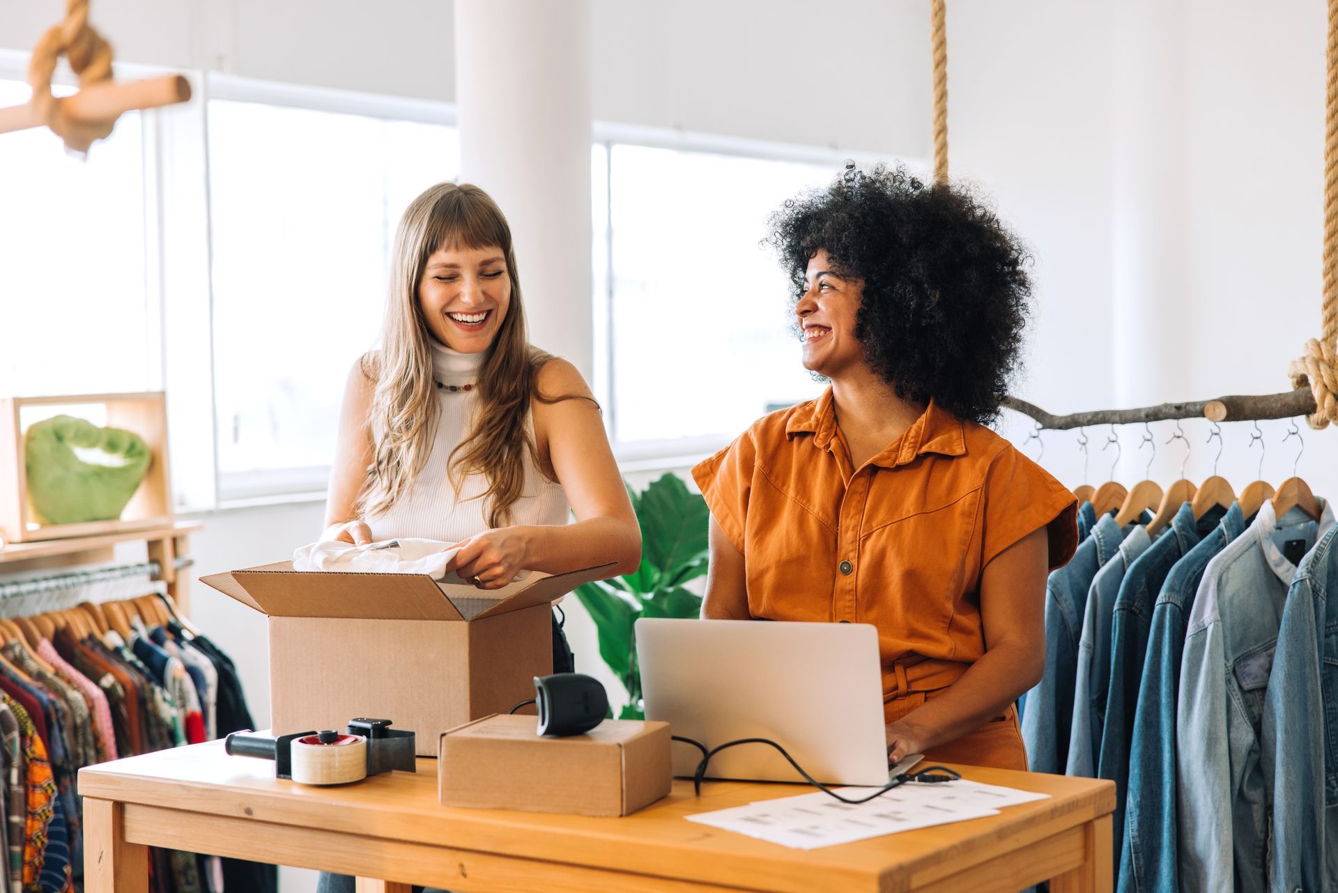 Two people packing a box in a clothing shop beside a laptop and rack of hanging clothes