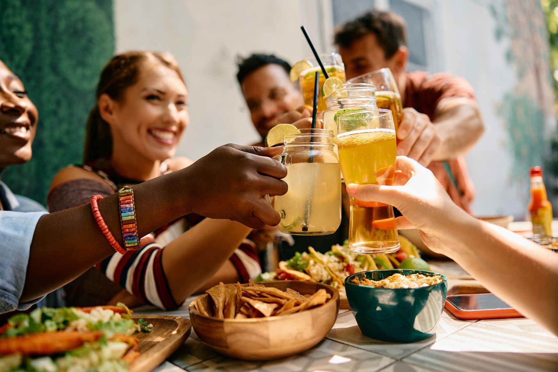 Friends clinking glasses over a colorful outdoor meal at a table with drinks and snacks