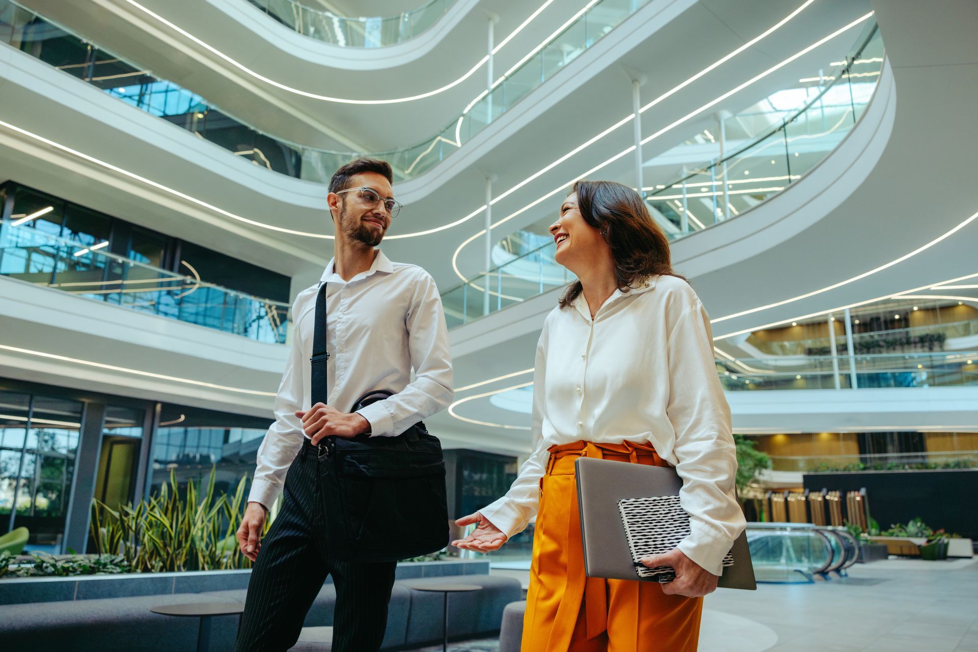 Two people walking in a modern atrium, one holding a laptop and wearing orange pants