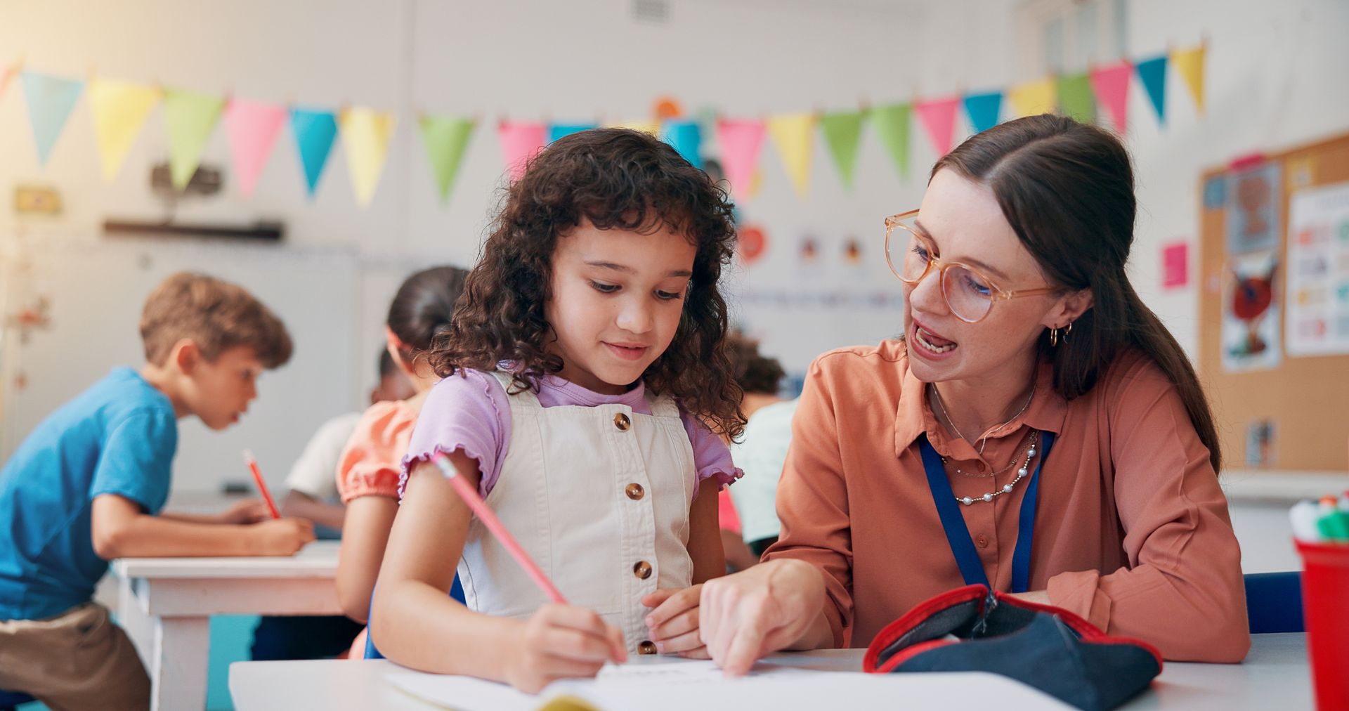 Teacher helping a child write at a classroom table during a craft activity, with colorful bunting in the background.