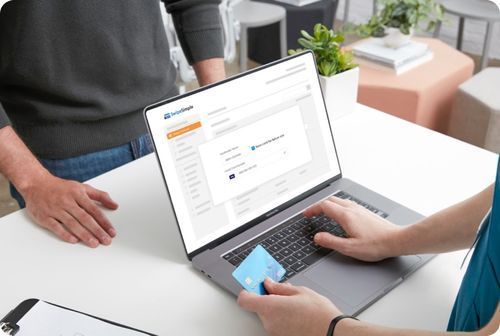Two people using a laptop at a white desk, with one holding a credit card in an online checkout screen