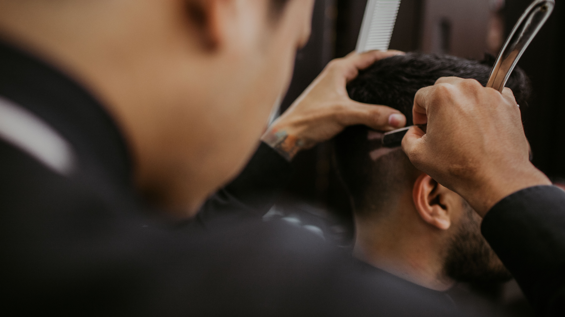 A barber uses a straight razor to groom the hairline of a client seated in a shop.