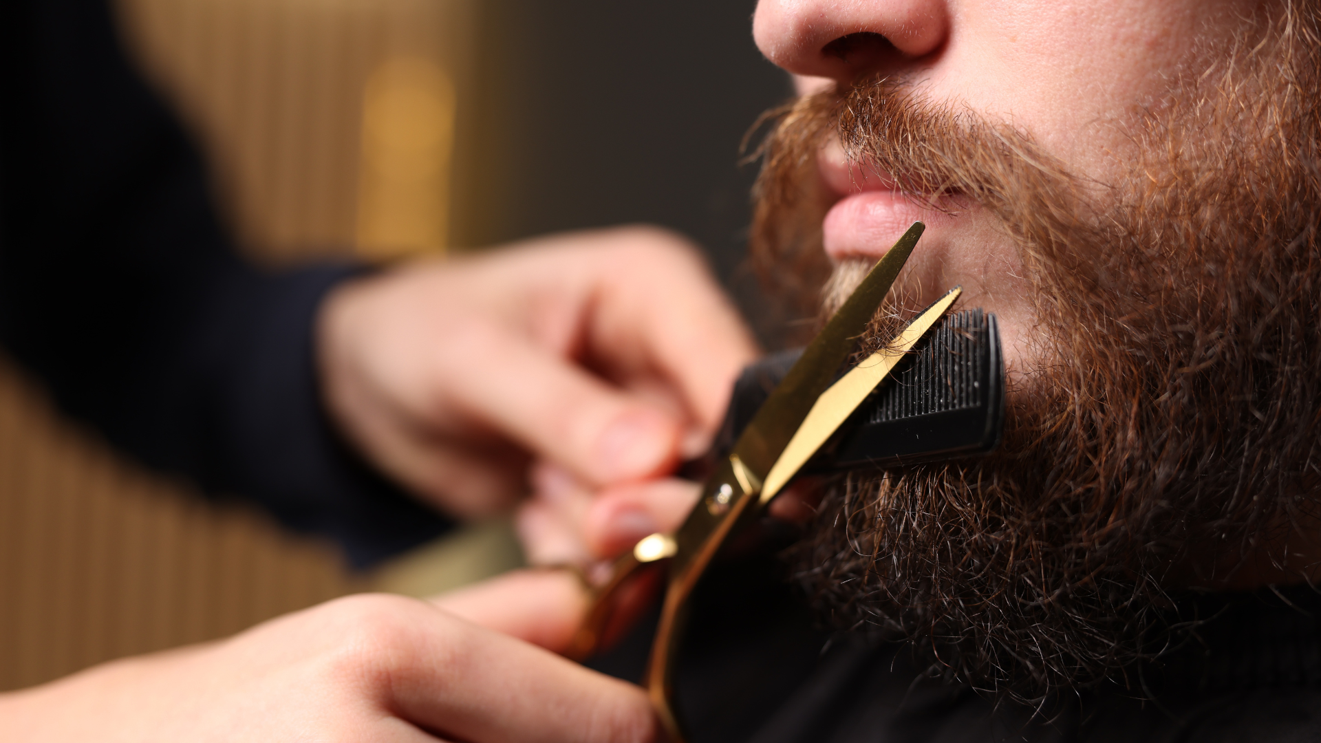 A barber uses gold scissors and a black comb to trim a person's thick, brown beard.