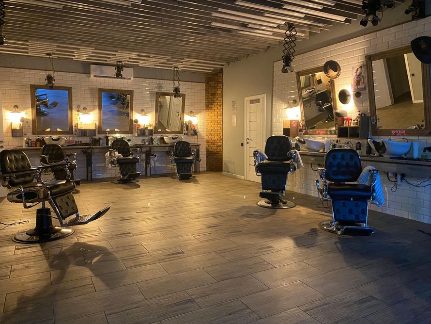A modern barbershop with five dark salon chairs, wooden workstations, and large mirrors against a white brick wall.
