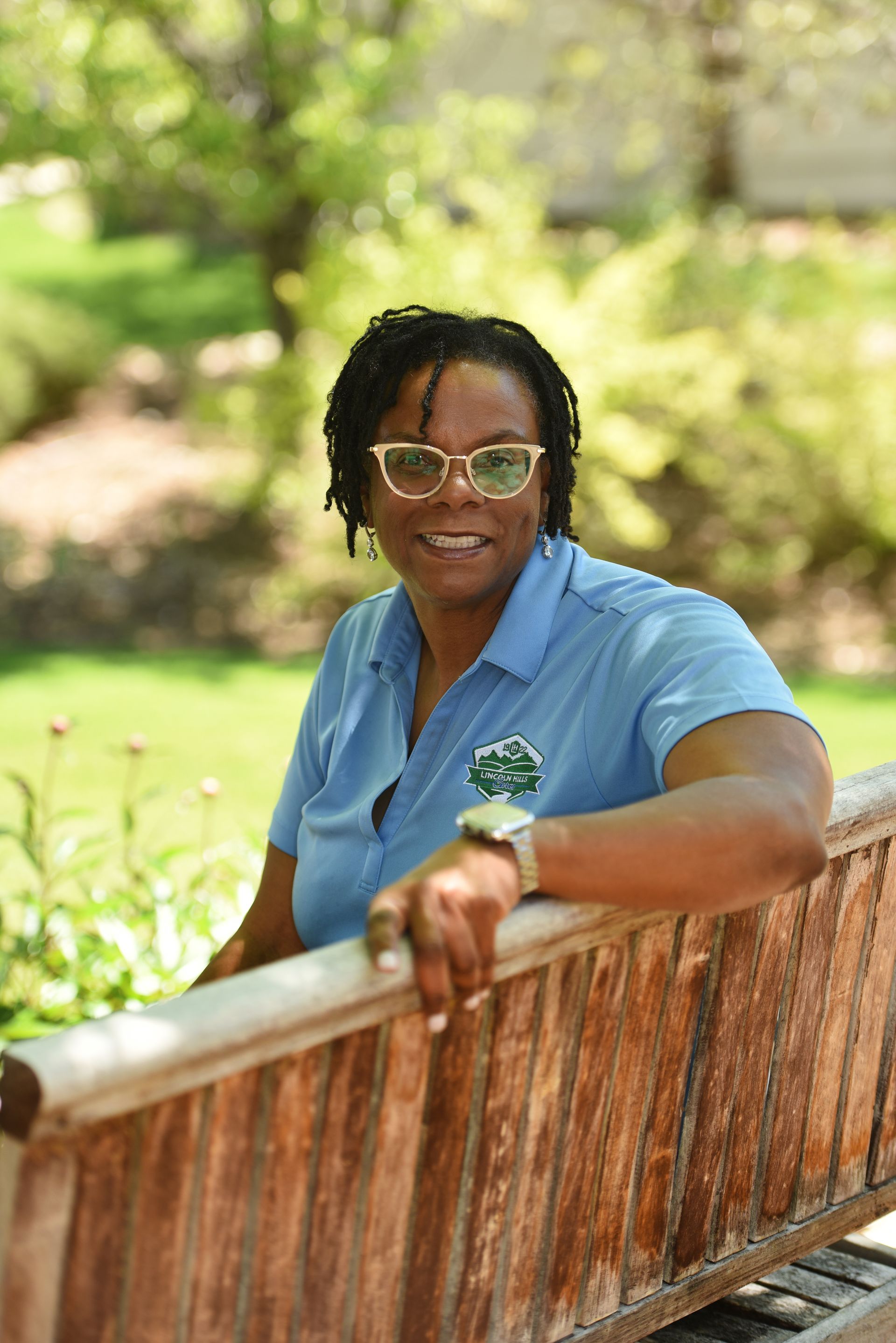 A woman in a blue shirt and glasses is leaning on a wooden bench.