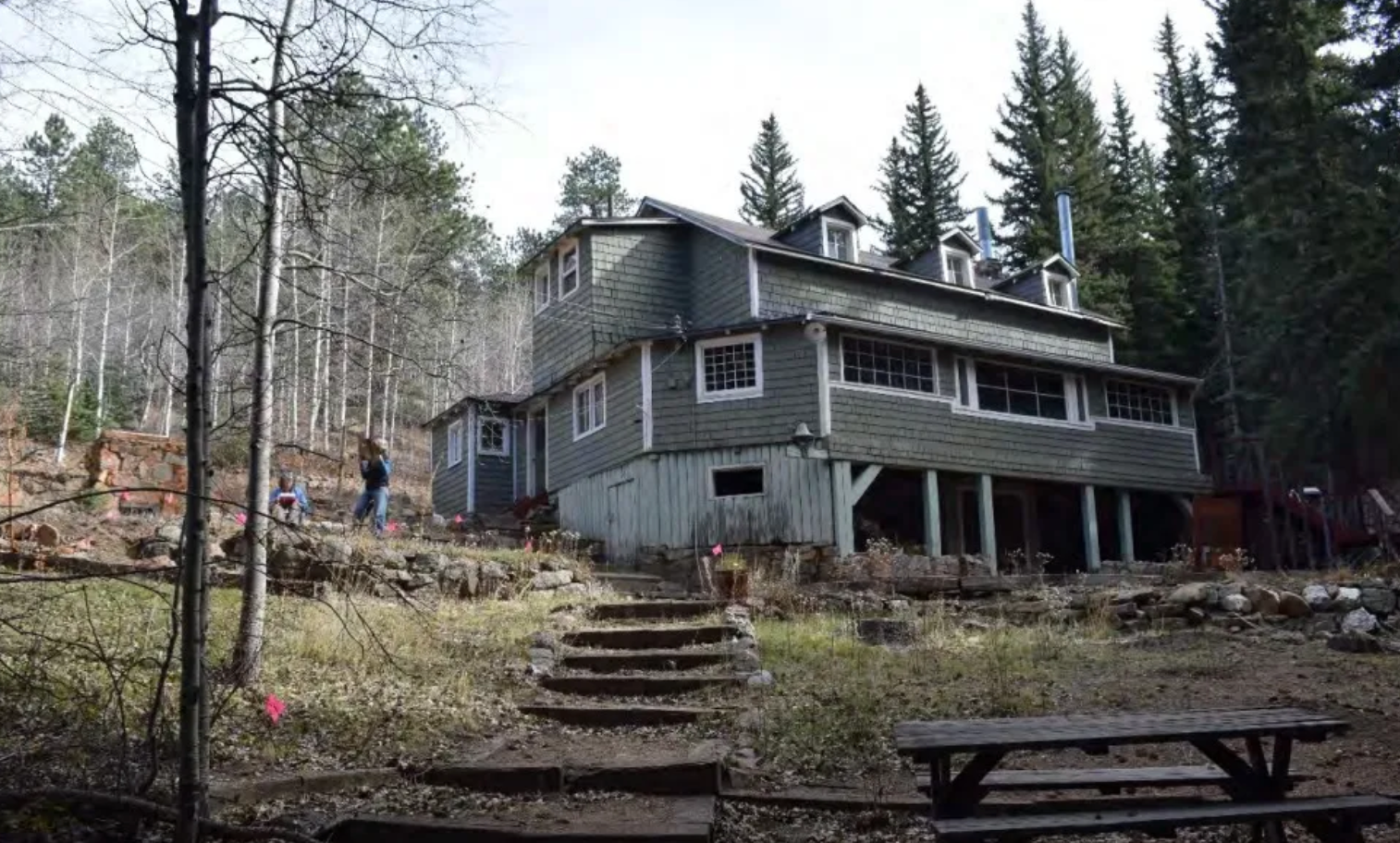 A large house in the middle of a forest with a picnic table in front of it.