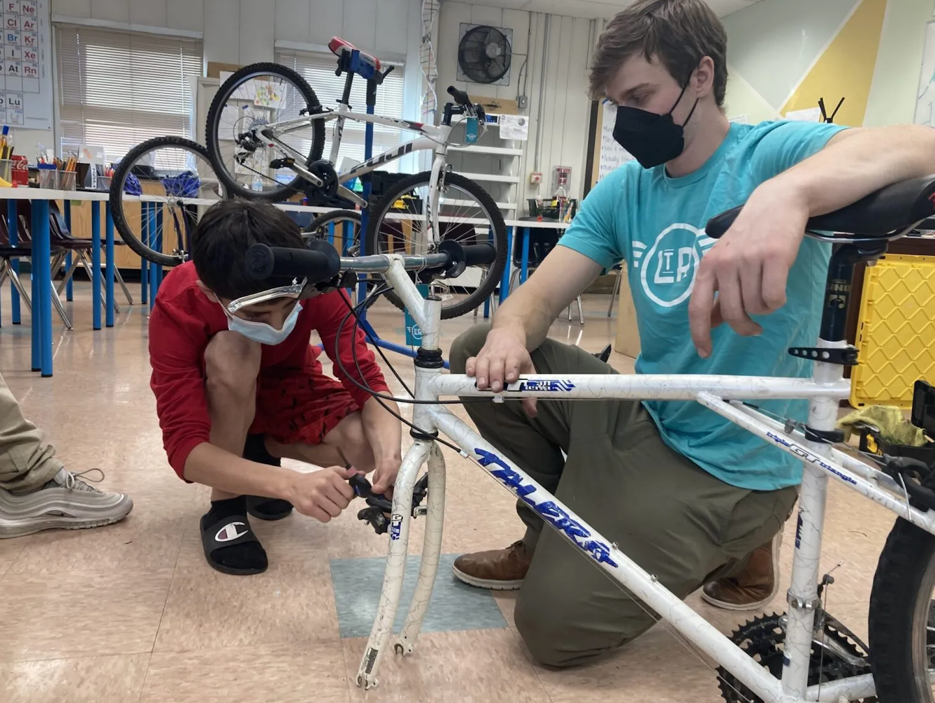 Two men wearing masks are working on a bicycle in a classroom.