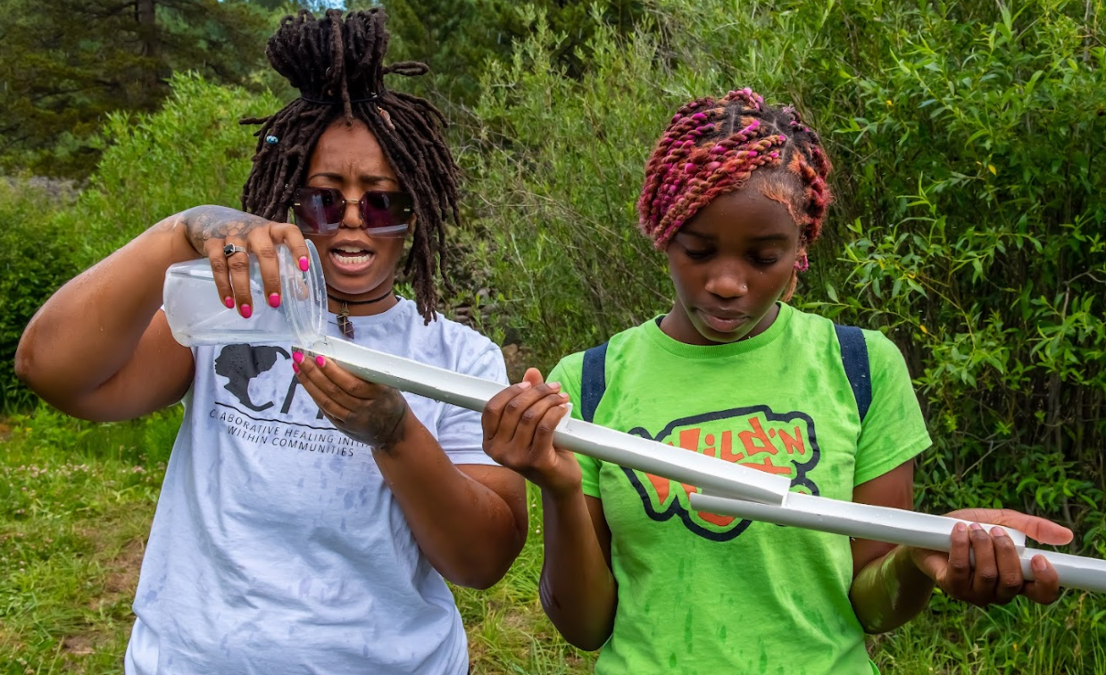 Two women are standing next to each other in a field holding a stick and a bottle of water.