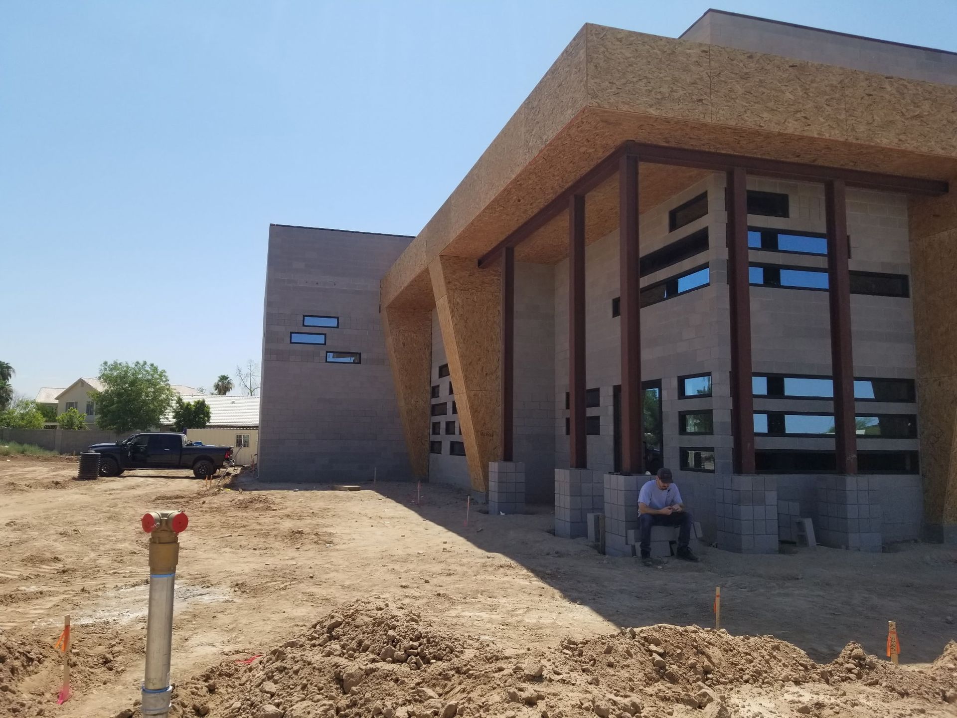 Modern building with tan brick and tall windows under construction on a dusty site; one person sits by the entrance.