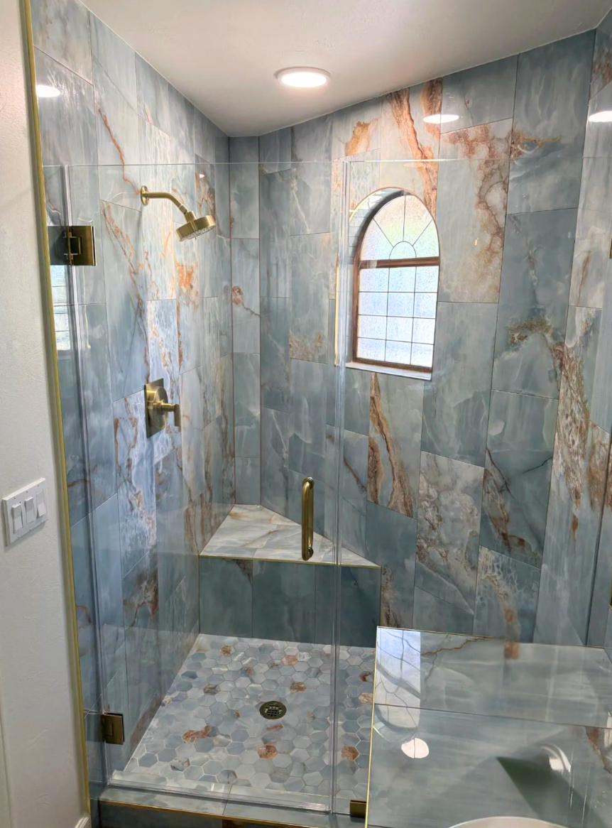 Blue and brown tiled shower with glass door and brass fixtures, small arched window.