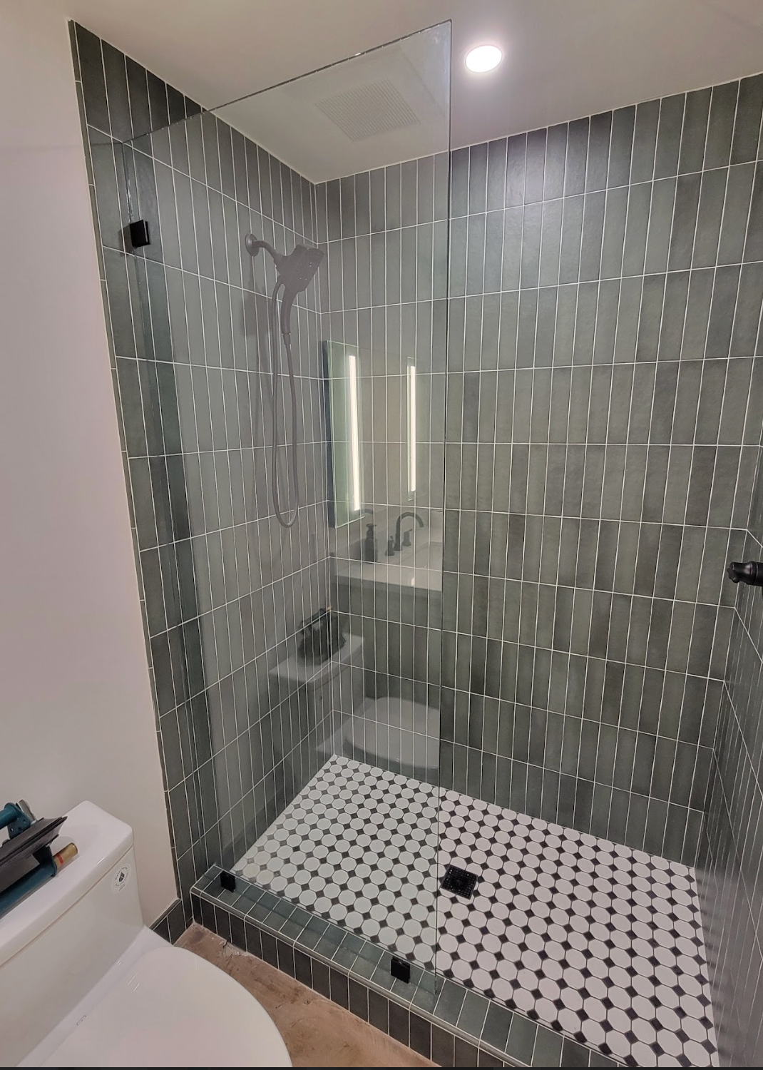 Modern shower with gray tiled walls, glass door, and black/white tiled floor.